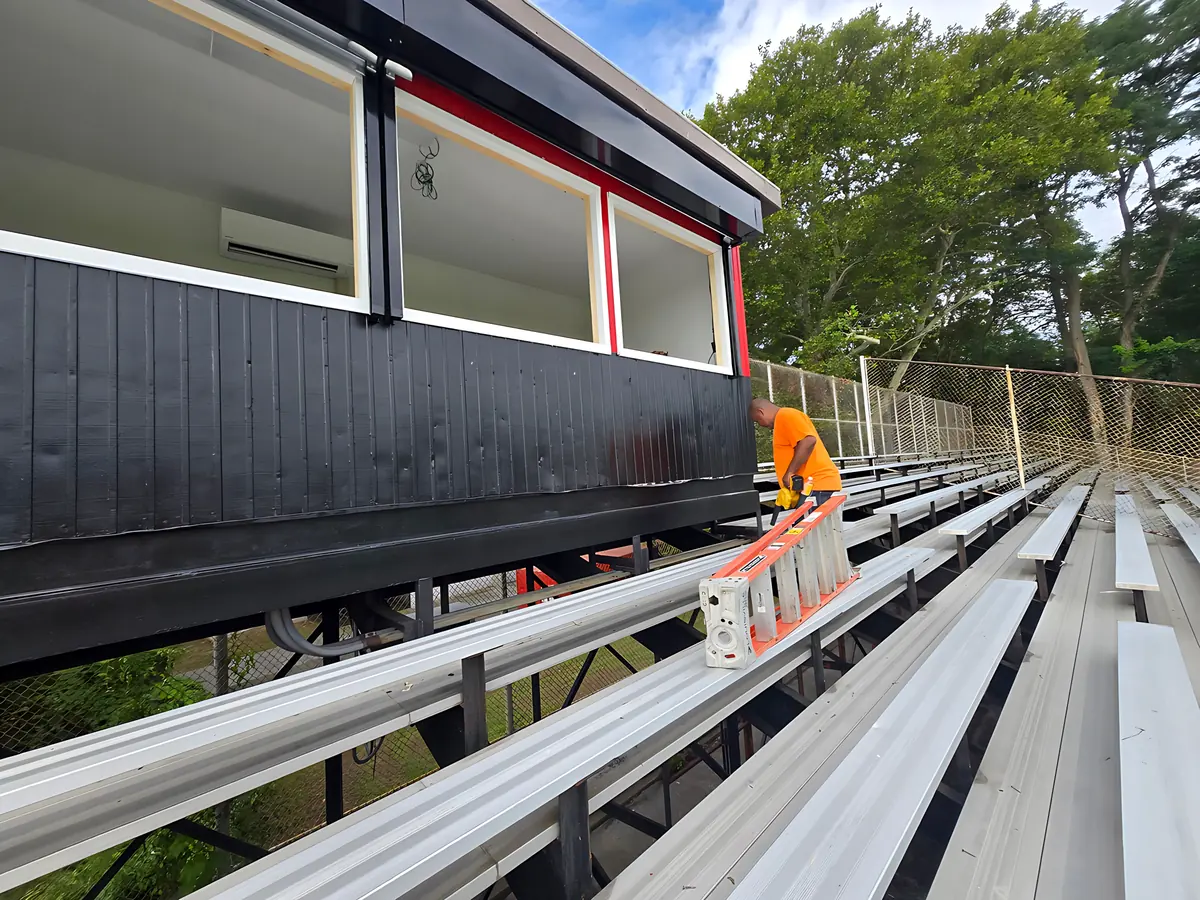 Crew member carrying a ladder along the bleachers beside a press box being reglazed